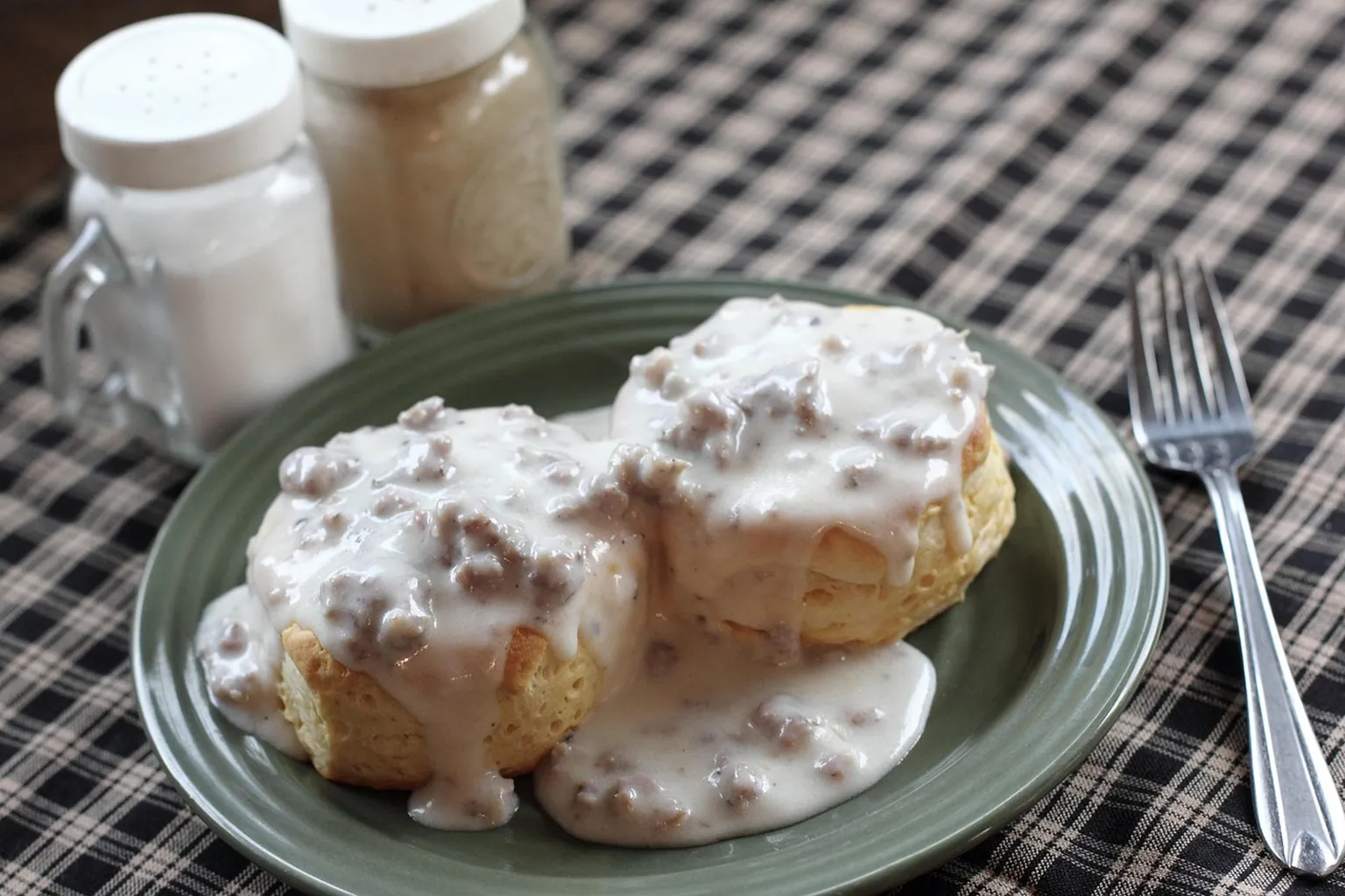 Two biscuits covered in sausage gravy on a green plate on a checked tablecloth